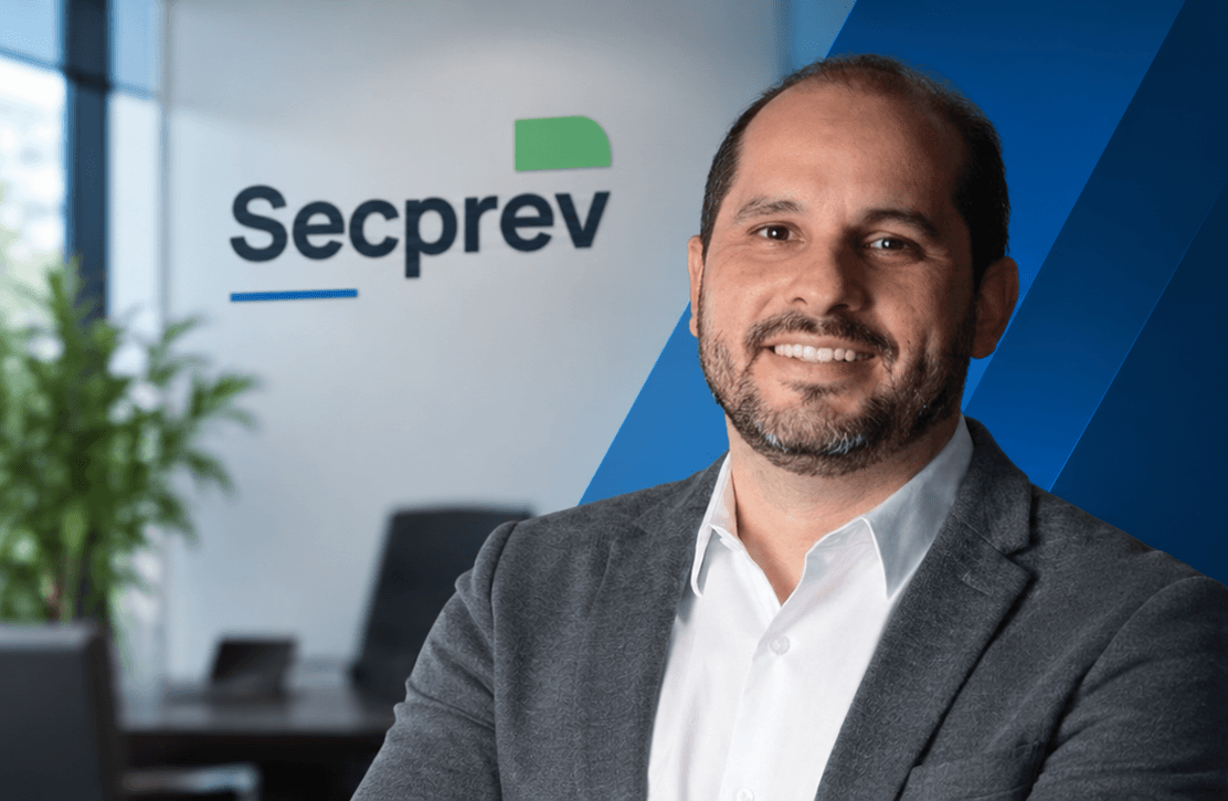 Heymard, consultor da Secprev, sorrindo em um escritório com o letreiro da Secprev ao fundo.